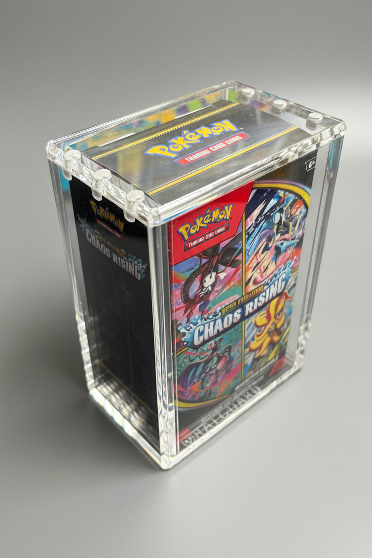 A clear acrylic display box containing a Pokémon TCG booster pack, photographed on a dark textured surface with colorful framed art in the background.