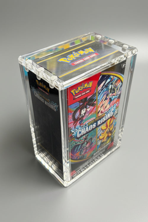 A clear acrylic display box containing a Pokémon TCG booster pack, photographed on a dark textured surface with colorful framed art in the background.