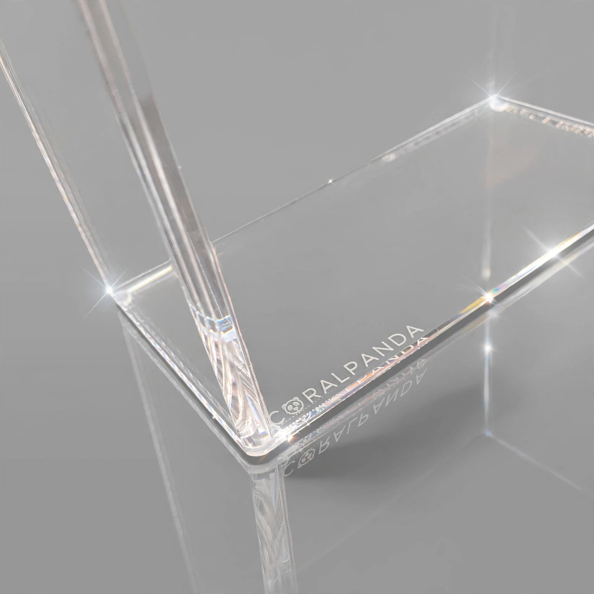 Clear acrylic rectangular display stand with corner brackets and a vertical support leg, featuring the brand name 'CORAL PANDA' printed on one edge, shown on a reflective gray surface.