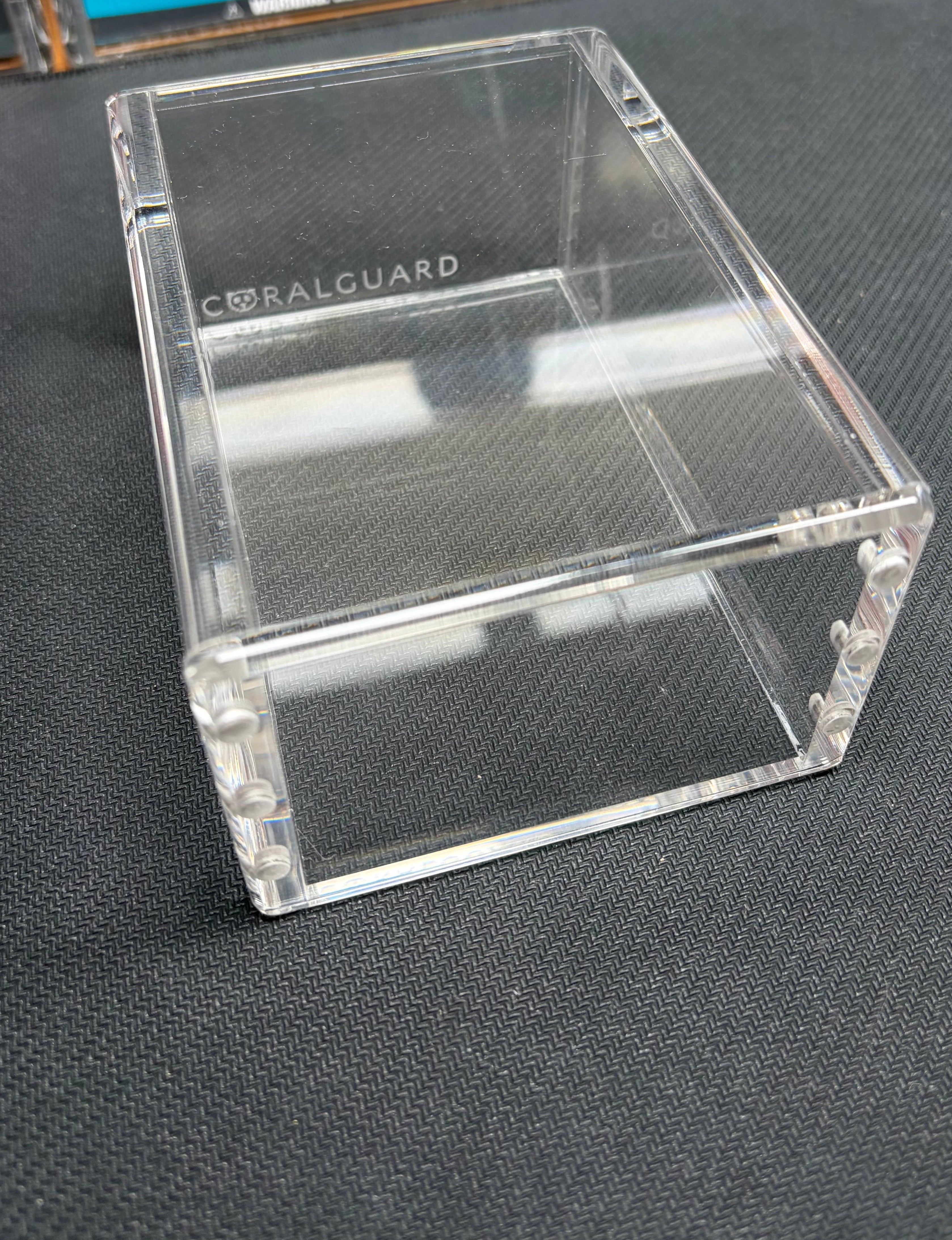 A clear acrylic display box with the brand name 'CORALGUARD' printed on the inside, resting on a dark textured surface.